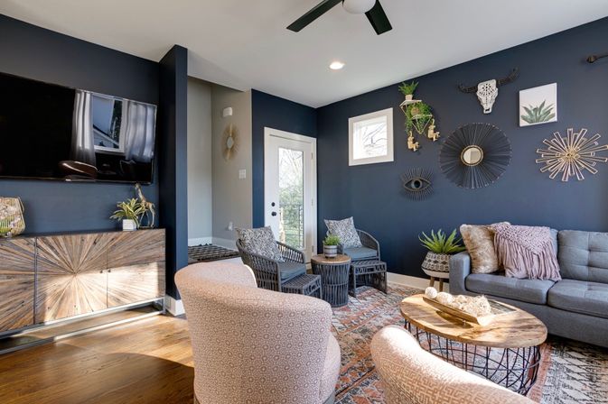 Cozy contemporary boho living room with a deep navy accent wall, gray sofa with blush throw pillows, two patterned blush chairs, round wooden coffee table, wicker side chairs, decorative sunburst wall art and hanging plants, hardwood floors and a door to a small balcony.