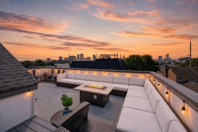 Rooftop patio with large white sectional sofa, central fire pit table, hanging Edison string lights and an urban city skyline at sunset