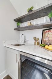 Bright modern wet bar with white marble countertop, stainless steel undermount sink and beverage fridge, dark floating wood shelves holding glassware and potted plants, framed print and citrus on a cutting board