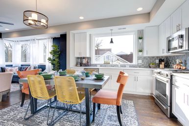 Sunny modern open-concept kitchen and dining area in a suburban home with white cabinets, stainless appliances, gray tile backsplash, wooden table with yellow and orange chairs, and a large front window