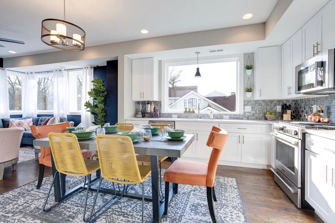 Sunny modern open-concept kitchen and dining area in a suburban home with white cabinets, stainless appliances, gray tile backsplash, wooden table with yellow and orange chairs, and a large front window