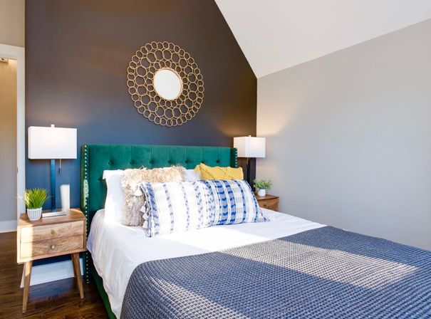 Bright modern bedroom with emerald green tufted headboard against a navy accent wall, circular gold wall mirror, white bedding with blue patterned lumbar pillow, gray textured throw, and wooden nightstands with lamps and potted plants.