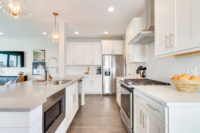Bright open-concept modern kitchen with white shaker cabinets, quartz countertops, stainless steel refrigerator and gas range, large island with sink and built-in microwave, pendant lights and hardwood floors