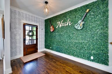 Sunlit home foyer with a dark wood front door, hardwood floors and geometric pendant light, plus a large faux boxwood feature wall displaying two mounted guitars and a white sign reading "Nash".