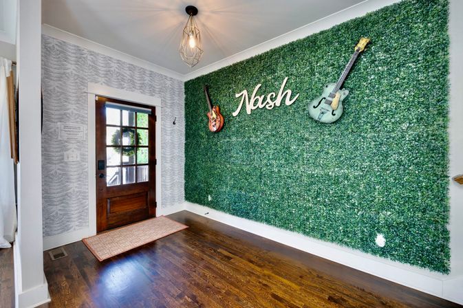 Sunlit home foyer with a dark wood front door, hardwood floors and geometric pendant light, plus a large faux boxwood feature wall displaying two mounted guitars and a white sign reading "Nash".
