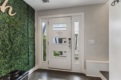 Modern foyer with a white front door featuring three horizontal glass panes and sidelights, faux boxwood accent wall, tufted black bench, coat hooks and dark hardwood floors.