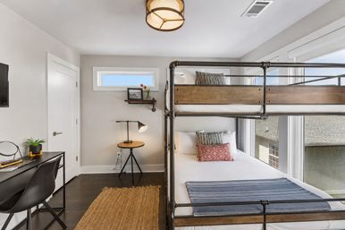 Bright modern bedroom with industrial metal-and-wood bunk bed, cozy lower bed with patterned pillows and blue throw, black study desk and chair with plant, round tripod side table and lamp, jute rug and large windows flooding the room with natural light.