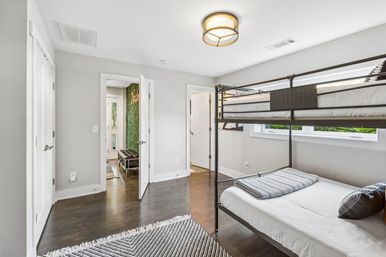 Bright contemporary bedroom with metal bunk bed, white and gray bedding, dark hardwood floors, geometric rug and gray walls, natural light from a wide window and an open doorway revealing a green living wall in the hallway.