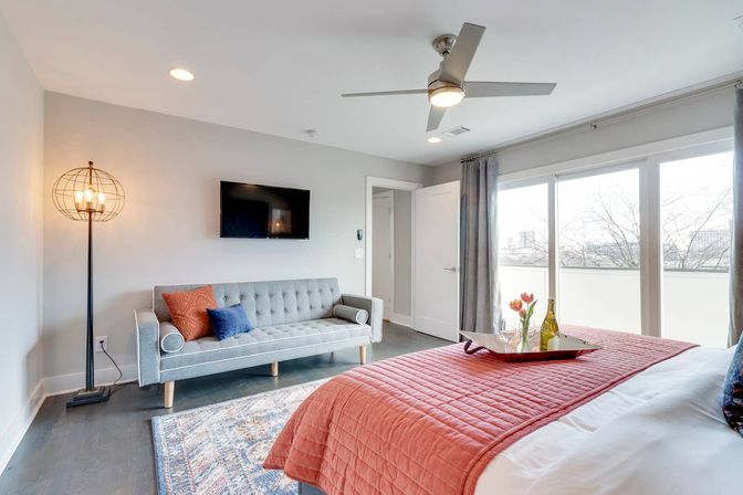 Bright modern urban apartment bedroom with coral quilted bed and tray of wine and tulips, gray tufted sofa with colorful pillows, wall TV, cage floor lamp, ceiling fan, and sliding glass doors to a balcony city view.