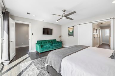 Bright sunlit modern bedroom with a large white bed and gray throw, emerald green velvet sofa, wall-mounted TV, brushed-metal ceiling fan, sliding barn door, dark hardwood floors and patterned area rug.