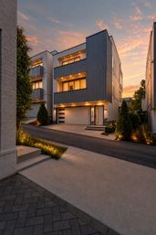 Contemporary three-story townhouse exterior with metal siding, illuminated balconies and garage, landscaped driveway and warm sunset sky in an urban neighborhood.