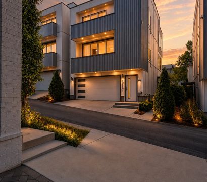 Contemporary three-story townhouse exterior with metal siding, illuminated balconies and garage, landscaped driveway and warm sunset sky in an urban neighborhood.