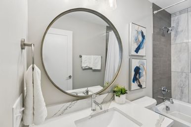 Modern bathroom interior with large round mirror over white marble countertop, chrome faucet, folded white towels, potted succulent, abstract blue artwork, and gray-tiled bathtub/shower.