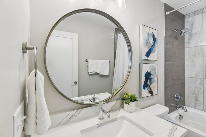 Modern bathroom interior with large round mirror over white marble countertop, chrome faucet, folded white towels, potted succulent, abstract blue artwork, and gray-tiled bathtub/shower.