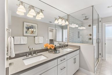 Bright modern master bathroom with double sinks on a gray quartz vanity, white cabinets, chrome faucets, large mirror, pendant lights, glass-enclosed hex-tile walk-in shower and peach roses on a decorative tray.