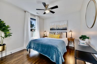 Bright modern bedroom with hardwood floors, ceiling fan, large window, blue throw blanket, yellow accent pillows, abstract wall art, bedside lamps, potted plant and round wall mirror