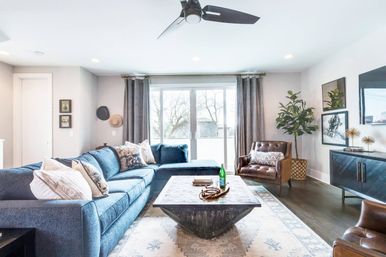 Bright modern living room with large sliding glass doors, blue L-shaped sectional with patterned pillows, geometric wooden coffee table on a pale patterned rug, brown leather armchair, potted plant, dark media console, and ceiling fan — cozy, airy seating area with natural light.