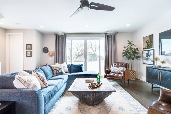 Bright modern living room with large sliding glass doors, blue L-shaped sectional with patterned pillows, geometric wooden coffee table on a pale patterned rug, brown leather armchair, potted plant, dark media console, and ceiling fan — cozy, airy seating area with natural light.