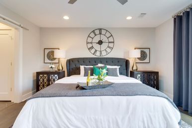 Inviting modern master bedroom with tufted gray headboard, oversized round wall clock, white bedding with gray throw and green accent pillows, matching nightstands and lamps, decorative tray with bottle and flowers, recessed lighting and dark curtains.