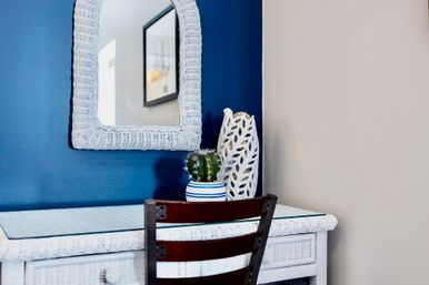 White wicker vanity and wicker-framed mirror on a navy blue accent wall, topped with a striped ceramic pot holding a small cactus and a sculptural white vase, with a dark wood chair in front.