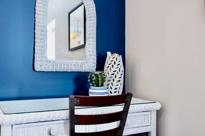 White wicker vanity and wicker-framed mirror on a navy blue accent wall, topped with a striped ceramic pot holding a small cactus and a sculptural white vase, with a dark wood chair in front.