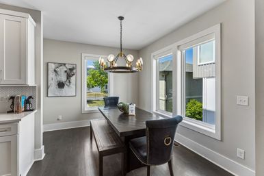 Sunlit modern dining nook with a dark wood farmhouse table and bench, navy velvet chairs with brass ring pulls, round brass chandelier, large windows, gray walls, hardwood floors and framed horse artwork.