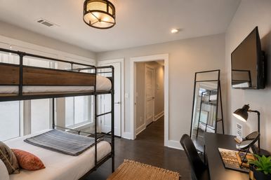Bright modern bedroom with industrial metal-and-wood bunk bed by large windows, wall-mounted TV, leaning full-length mirror, black desk with lamp and chair, hardwood floors and woven rug — cozy, stylish layout.