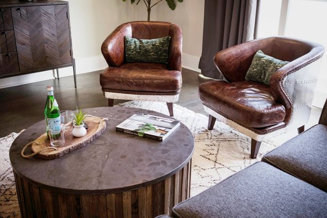 Cozy modern rustic living room interior with two brown leather accent chairs with green pillows, round wooden coffee table with a glass bottle, tray and book on a patterned rug.