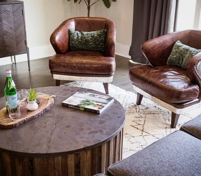 Cozy modern rustic living room interior with two brown leather accent chairs with green pillows, round wooden coffee table with a glass bottle, tray and book on a patterned rug.