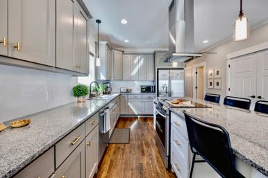 Bright modern kitchen with gray cabinets, white subway tile, granite countertops and a large island featuring a stainless steel range and refrigerator, hardwood floors, pendant lights and black bar stools.