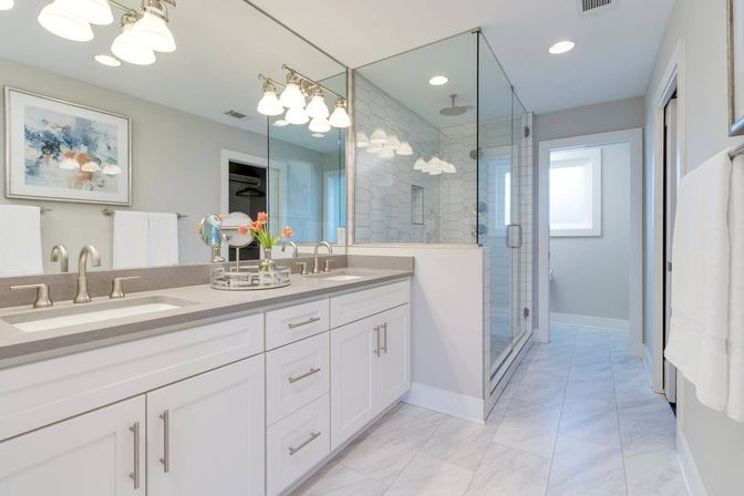 Bright, spa-like modern bathroom with white double-sink vanity, gray quartz countertop, large mirror and pendant lights, glass-enclosed walk-in shower with subway tile, marble-look floor tiles and fresh tulips on the counter.