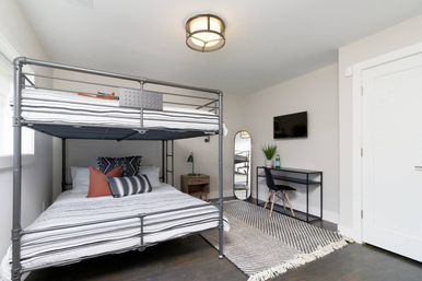 Bright modern bedroom interior with gray metal bunk bed and striped linens, decorative pillows, woven rug, small black desk and chair, wall-mounted TV, full-length mirror and bedside table — cozy minimalist guest room.