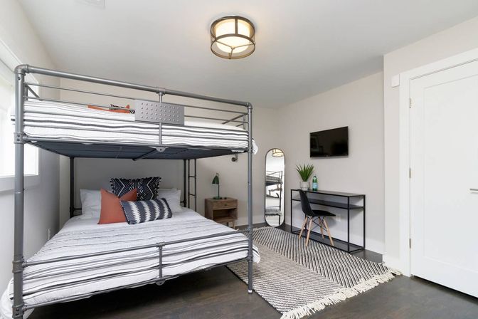 Bright modern bedroom interior with gray metal bunk bed and striped linens, decorative pillows, woven rug, small black desk and chair, wall-mounted TV, full-length mirror and bedside table — cozy minimalist guest room.