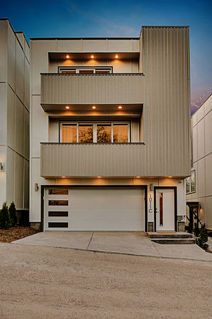 Contemporary three-story townhouse with stacked balconies, warm recessed lights, white two-car garage and driveway at dusk