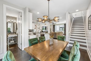 Bright open-concept dining area in a modern home with a reclaimed wood table, green velvet chairs, industrial chandelier, adjacent white kitchen, staircase, and patterned powder room.
