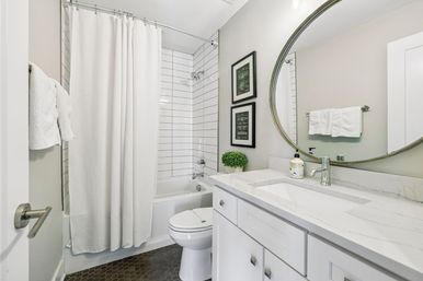Bright contemporary small bathroom with white tub/shower and subway tile, white shower curtain, round mirror above marble-look vanity, white cabinets, black hexagon floor tiles, framed art and small potted plant.