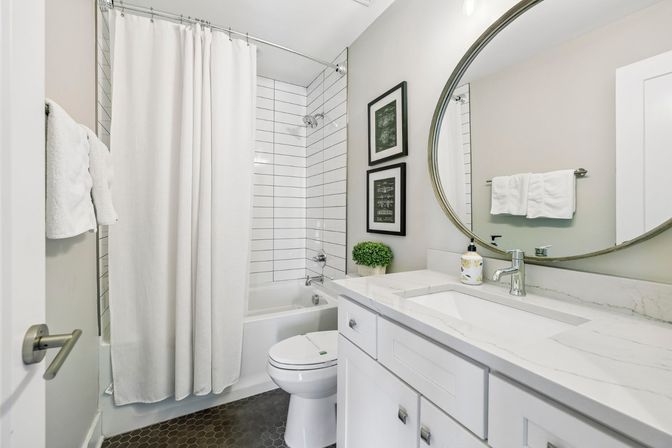 Bright contemporary small bathroom with white tub/shower and subway tile, white shower curtain, round mirror above marble-look vanity, white cabinets, black hexagon floor tiles, framed art and small potted plant.
