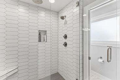 Bright modern walk-in shower tiled in elongated white hexagons with a glass door, brushed-nickel shower fixtures and a recessed niche stocked with toiletries.
