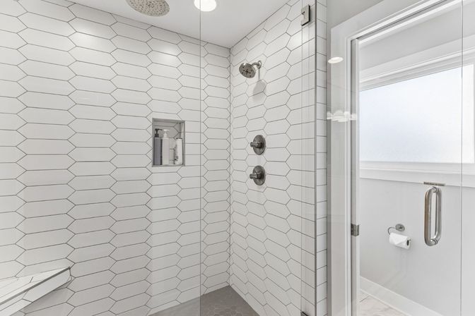 Bright modern walk-in shower tiled in elongated white hexagons with a glass door, brushed-nickel shower fixtures and a recessed niche stocked with toiletries.