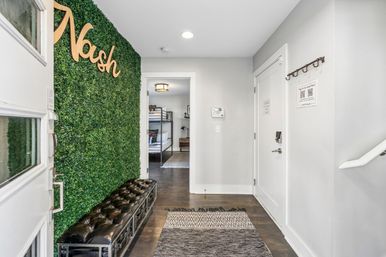 Modern entryway with green boxwood accent wall and wooden script sign, tufted black leather bench, dark hardwood floors, patterned rug and doorway to a bunk-bed bedroom.