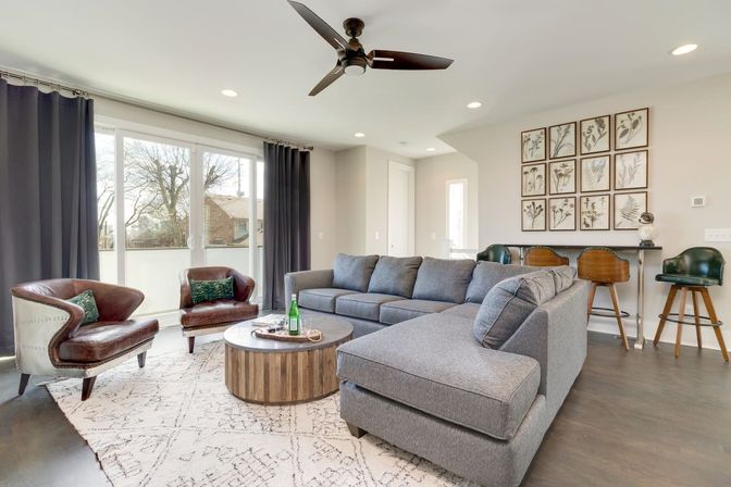 Bright contemporary living room with a gray sectional sofa, two brown leather armchairs, round wooden coffee table, sliding glass doors and ceiling fan