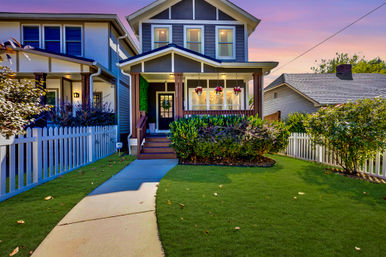 Two-story craftsman-style suburban house with a covered front porch, hanging flower baskets, white picket fence, manicured green lawn and curved concrete walkway under a colorful sunset sky