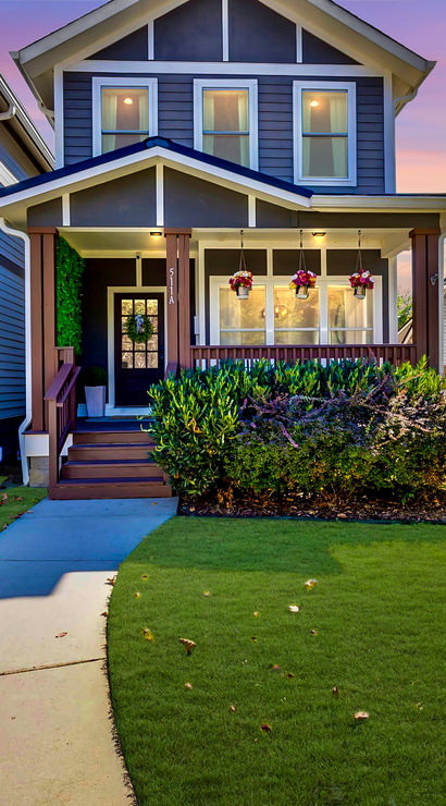 Two-story craftsman-style suburban house with a covered front porch, hanging flower baskets, white picket fence, manicured green lawn and curved concrete walkway under a colorful sunset sky