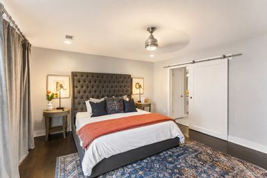 Modern master bedroom interior with tall gray tufted headboard, white bedding and rust-orange throw, patterned blue area rug, bedside tables with lamps and tulips, sliding barn door and ceiling fan
