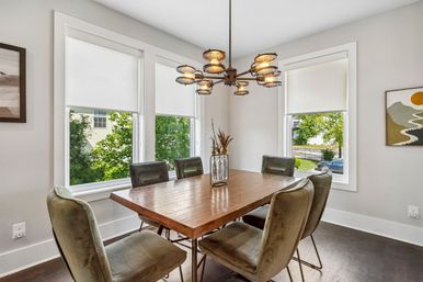 Sunlit residential dining room with a rustic wooden table, six olive-green velvet chairs, a vintage-industrial multi-light chandelier, and three large windows with white roller shades.