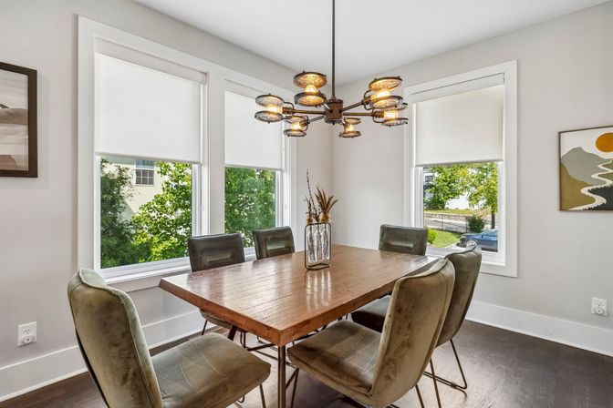 Sunlit residential dining room with a rustic wooden table, six olive-green velvet chairs, a vintage-industrial multi-light chandelier, and three large windows with white roller shades.