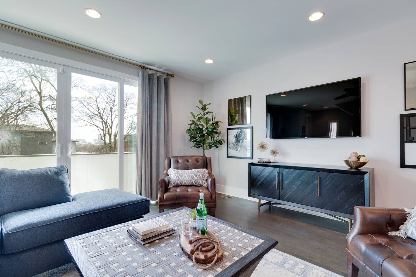 Sunlit contemporary living room with blue sectional, brown leather armchairs, woven coffee table with books and a glass bottle, wall-mounted TV over a dark wood console, potted plant, and large sliding glass doors opening to a tree-lined balcony view.