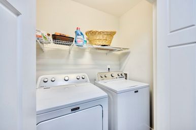 Bright home laundry closet with side-by-side top-loading washer and dryer, wire shelf above holding laundry detergent, small storage baskets and a woven laundry basket — neat and ready for laundry day.