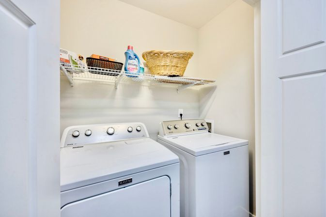 Bright home laundry closet with side-by-side top-loading washer and dryer, wire shelf above holding laundry detergent, small storage baskets and a woven laundry basket — neat and ready for laundry day.