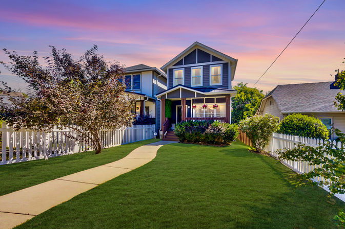 Charming two-story suburban house with covered front porch, hanging flower baskets, white picket fence, curving sidewalk and manicured front yard under a colorful sunset sky.